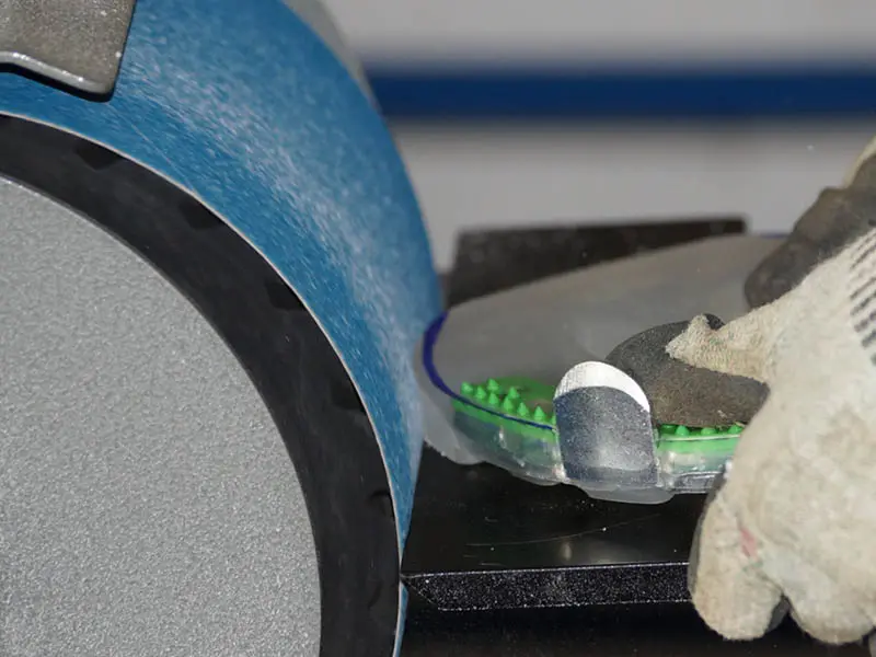Adjusting a horseshoe with a metal core and shock-absorbing plastic outer layer on a belt grinder. The horseshoe features an additional steel edge in the toe area. The farrier is wearing cut-resistant gloves.