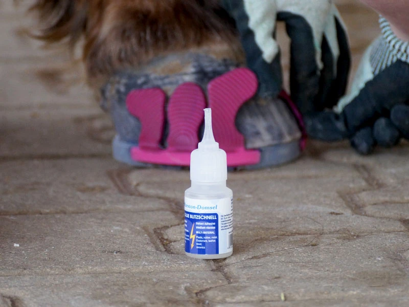The MD-Glue Blitzschnell by Marston Domsel is standing on paving stones; in the background, the farrier is gluing pink Wolf Busch® adhesive tabs onto the horse’s front hoof.