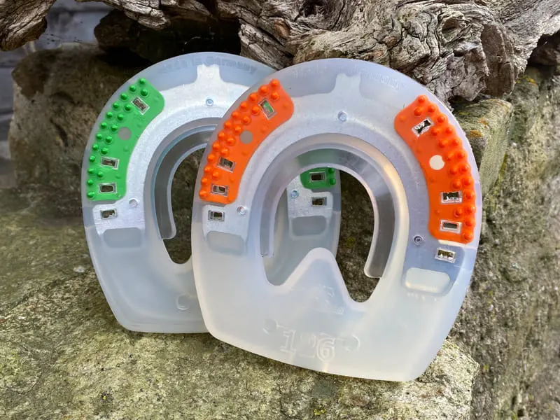 Two un-clipped composite horseshoes for front hooves, with green and orange textured inserts, placed side by side on stone
