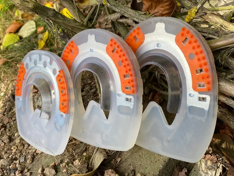 Three shapes of composite horseshoes without quarter clips and with orange textured inserts, leaning against a root on stone