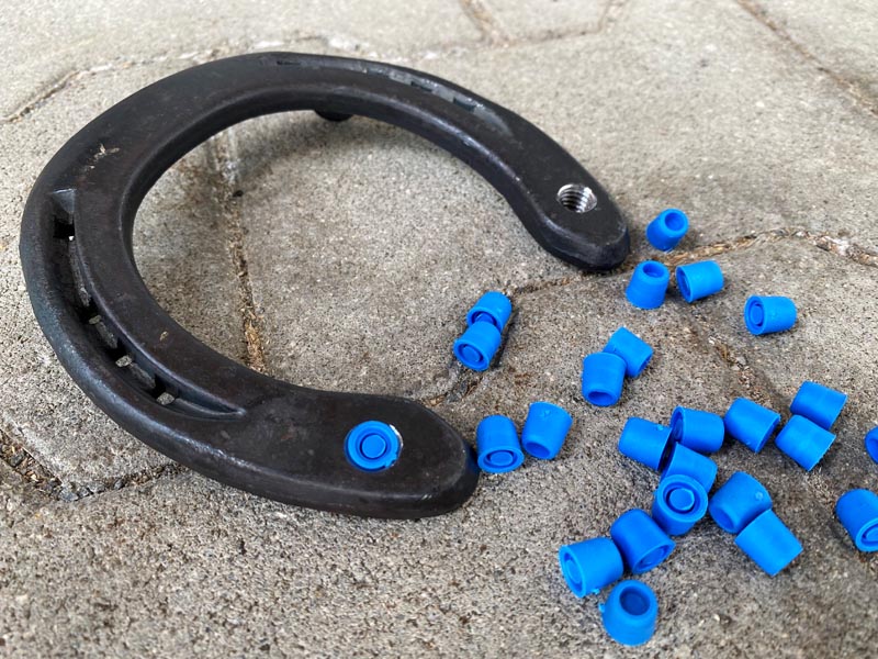 blue closure plugs are placed next to a steel horseshoe with stud holes; one stud hole is already sealed with a blue closure plug