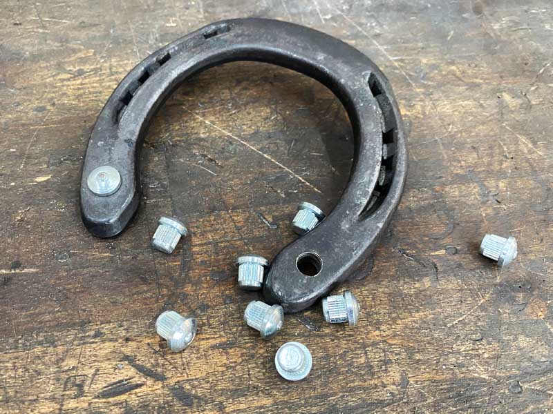 Beside a steel horseshoe, there are traction studs on a wooden floor. The horseshoe has two threaded holes, and one already has a drive-in stud mounted