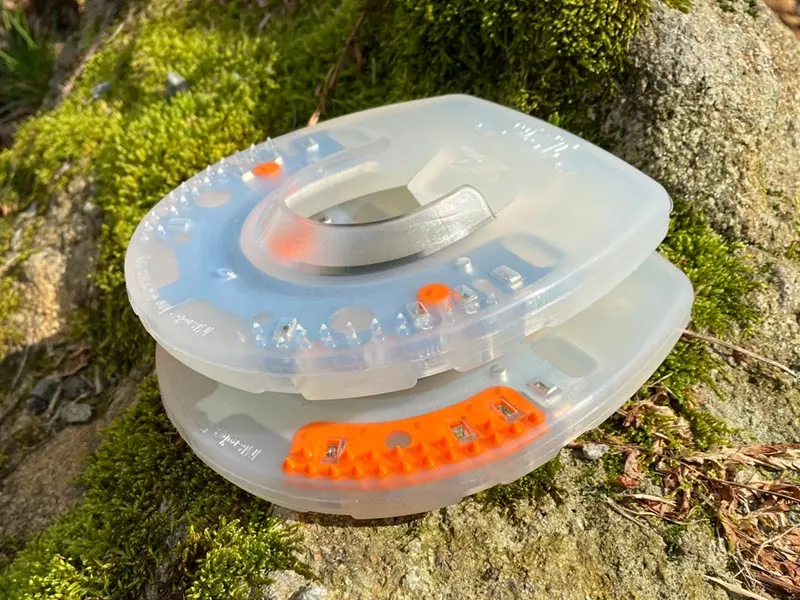 Two urethane-coated horseshoes without clips placed on a moss-covered stone. The top shoe is a Duplo Nailless model without a rigid stud insert, while the lower one is a standard model with an orange stud insert.