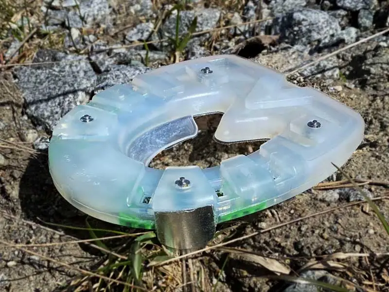 plastic horseshoe for higher temperatures, identifiable by green stud inserts, lying on a gravel path; the horseshoe is equipped with four spikes similar to the pins on a metal shoe