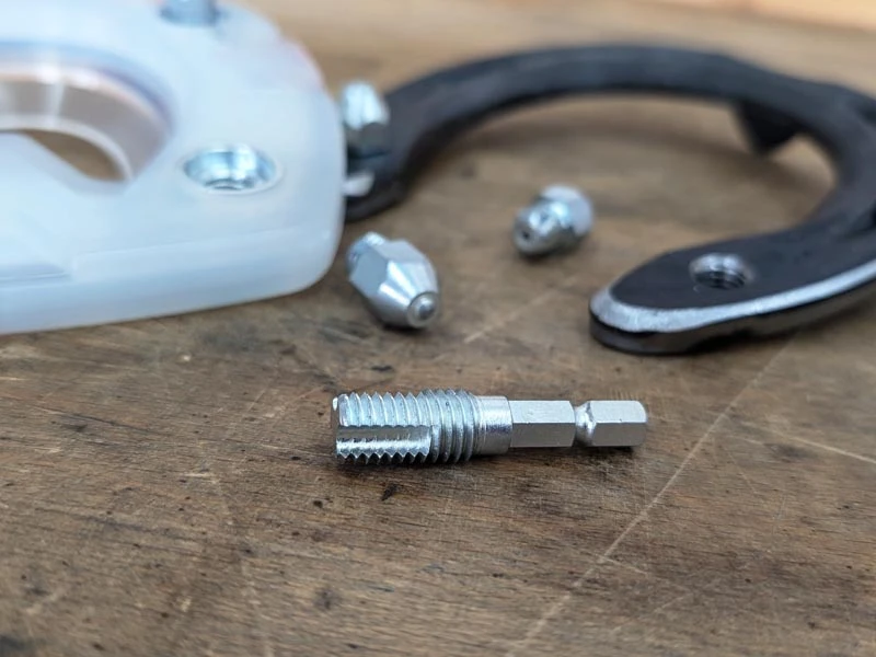 Un embout de nettoyage de filetage pour nettoyer le trou de crampon est posé sur un établi. En arrière-plan, on voit des ferrures avec des trous de crampons.