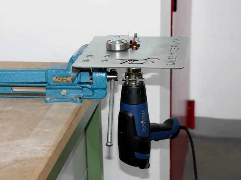 una pistola de aire caliente de la marca Steinel está sujeta a un dispositivo para la soldadura de herraduras adhesivas, que a su vez está fijado en un tornillo de banco de color azul claro.