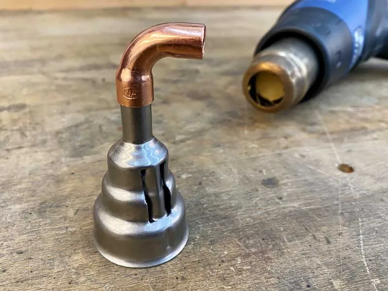 una boquilla reductora con extensión especial de cobre está sobre una mesa de trabajo, en el fondo hay una pistola de aire caliente azul