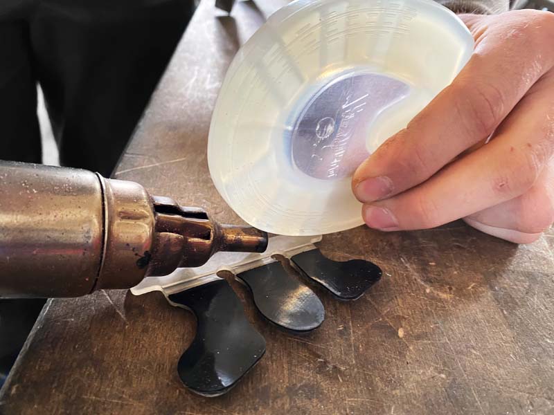 a farrier holds a pony horseshoe while welding it to an adhesive tab using a hot air gun with a reduction nozzle, the adhesive tab is placed on a workbench.